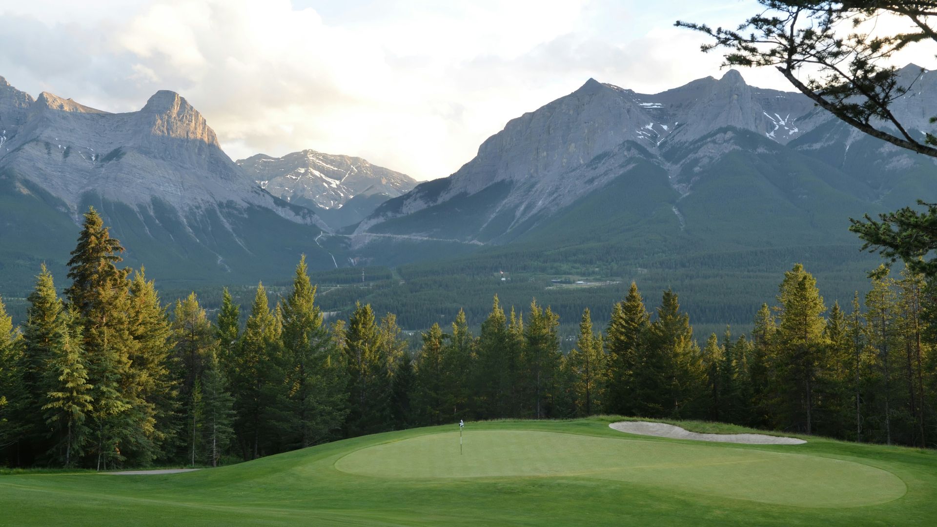 A scenic landscape print showing a golf course green and sand bunker bordered by a dense pine forest, with majestic mountains rising in the background under a cloudy sky.