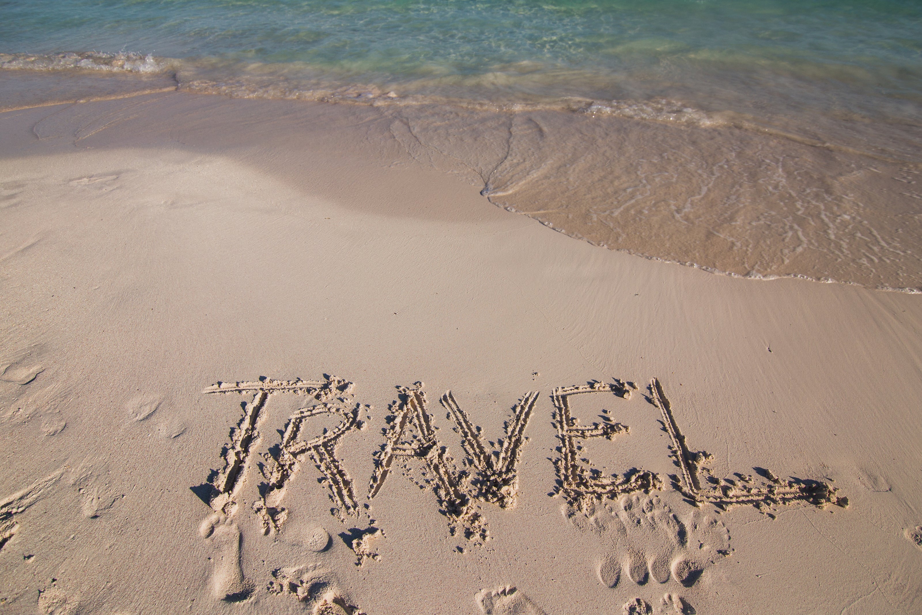 The word "TRAVEL" written in sand on a beach with turquoise waves washing ashore.